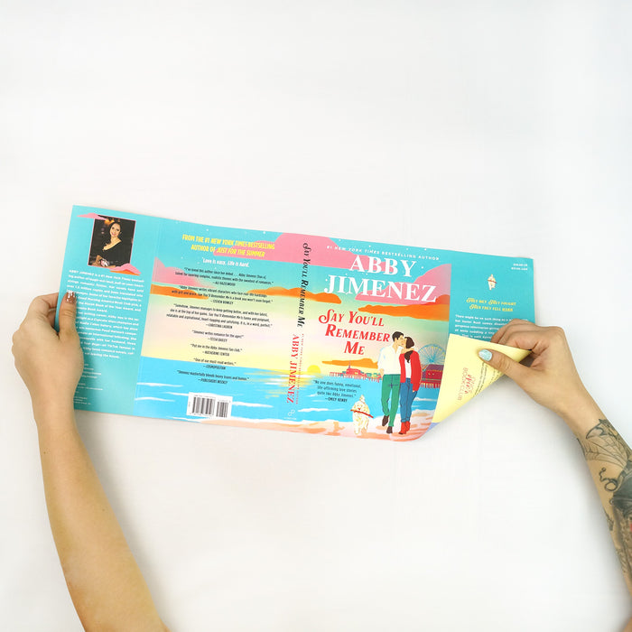 A pair of hands showing the first side of the reversible dust jacket for Say You'll Remember Me by Abby Jimenez featuring a couple walking on a beach with a pier and ferris wheel in the distance. This is the standard artwork from the original edition of Say You'll Remember Me.