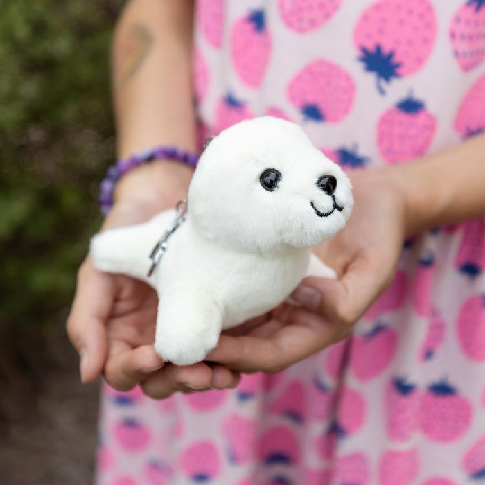 A white plush seal on a keychain is held in two hands in front of a girl wearing a strawberry-patterned dress.