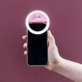 A pink ring light that says Smile! attached to the top of an iPhone held out in front of a purple background