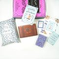 A custom, hardcover edition of Sisters of Fortune by Anna Lee Huber exclusively designed by Once Upon a Book Club sits on top of a pink Once Upon a Book Club Adult box. Surrounding this are three wrapped gifts labeled with page numbers, a signed bookplate, custom bookmark, art print, and book club kit.