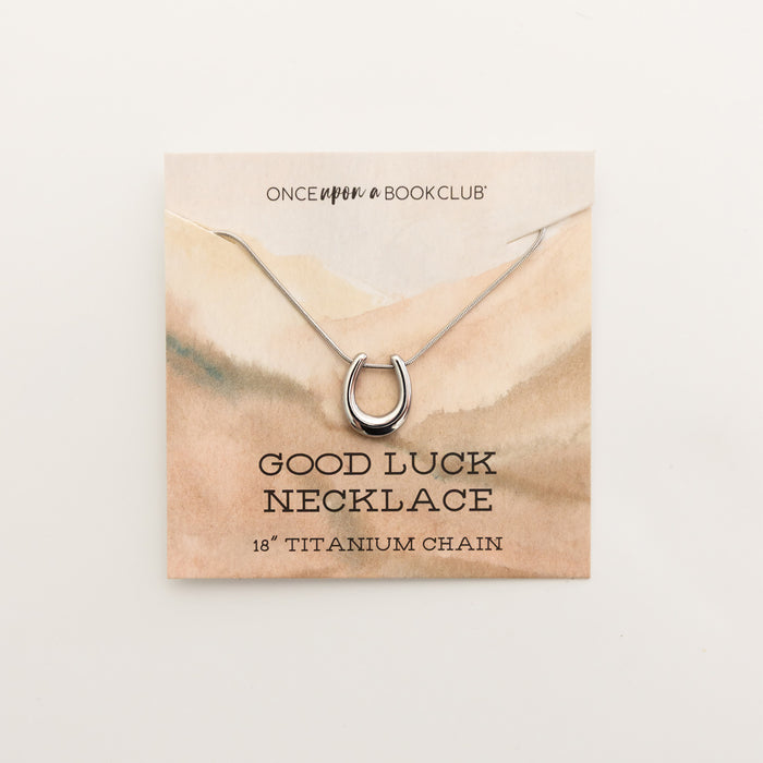 A silver horseshoe necklace on a backing card. The backing card says Once Upon a Book Club Good Luck Necklace 18" Titanium Chain.