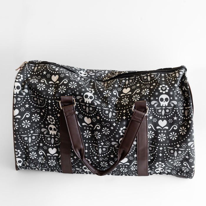 A black duffel bag with dark brown straps. The duffel bag has a pattern of white skulls, hearts, and stars on it.