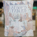 a person holds up a pastel-toned blanket labeled "Aesop's Fables"