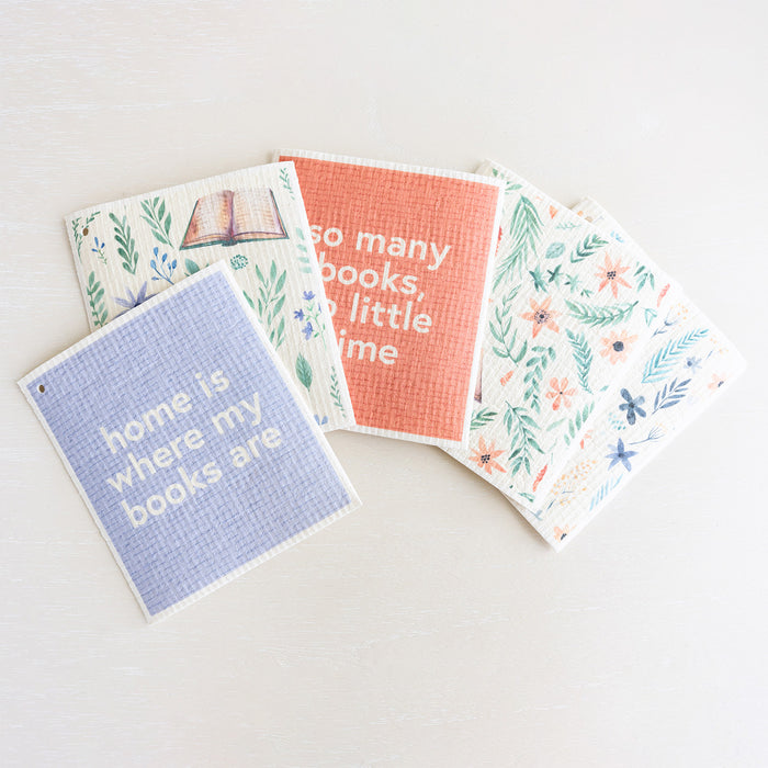 A collection of Swedish dishcloths in pastel tones featuring flowers and books. The top towel reads "home is where my books are"