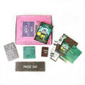 Paperback edition of The Family Inside lays on a pink OUABC box. In front of the box are four themed wrapped gifts labeled with page numbers and paper items.