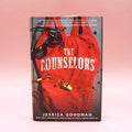 A hardcover copy of The Counselors by Jessica Goodman on a pink background.