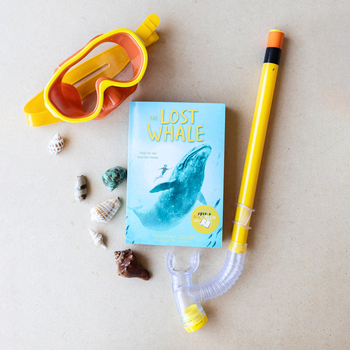 A paperback copy of The Lost Whale by Hannah Gold sits on a tan background. Surrounded by sea shells, yellow and orange diving goggles, and a snorkle.