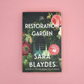 Book cover of 'The Restoration Garden' by Sara Blaydes on a pink background