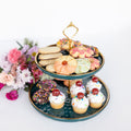 A two-tier dark green cake stand with gold accents filled with cupcakes and cookies on each level. A bouquet of flowers is laying behind the cake stand.