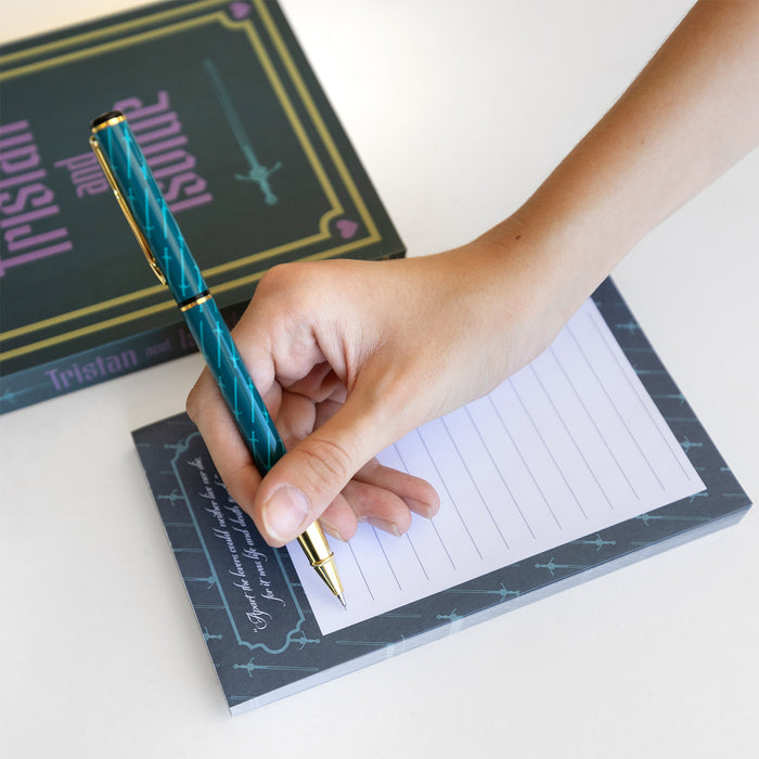 A hand holds a gold and blue pen with a light blue sword pattern on it over a lined notepad with matching background. Next to the notepad is a box labeled Tristan and Isolde.
