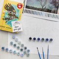 A paint by number kit with a large white paper, multiple sets of paints, reference image of a moonlit tree, 3 paintbrushes, and a paperback edition of Improbably Yours with an open your gift sticker on the front