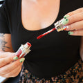 A woman with St. Patrick's Day themed nails holds a red lipstick tube with the wand out, showing a red color.