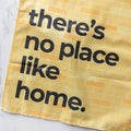 Yellow tea towel with black text "there's no place like home" on a brick-patterned background.